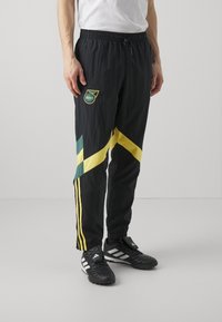 Pantalones de chándal negros con rayas diagonales amarillas y verdes, cinturilla elástica y detalle del logo en el muslo izquierdo. Tela ligera y texturizada.