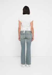 Λευκό τοπ με κοντά μανίκια και δέσιμο στην πλάτη· συνδυασμένο με ανοιχτό μπλε τζιν καμπάνα. Το μοντέλο φοράει λευκά παπούτσια και είναι γυρισμένο με την πλάτη στην κάμερα.