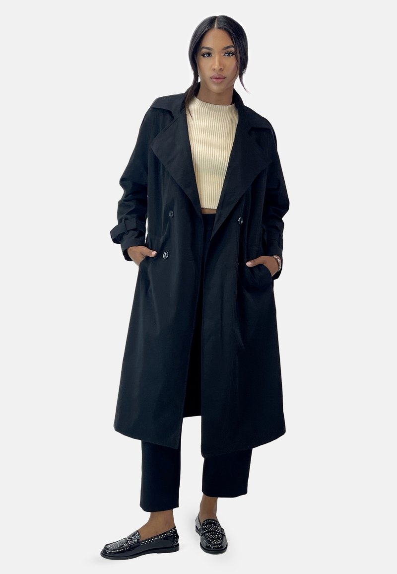 Abrigo negro de trench con un amplio cuello, diseño de doble botonadura y grandes bolsillos laterales, combinado con una blusa beige de canalé y pantalones negros.