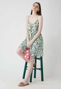 Haljina s cvjetnim uzorkom u zelenoj i krem boji, s trakama na ramenima i V-izrezom. Model drži ružičastu i krem beaded torbicu, nosi smeđe sandale.
