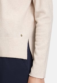 Helle beige Strickjacke mit strukturierter Oberfläche, geripptem Saum, seitlichen Schlitzen und einem kleinen metallischen Akzent nahe dem Saum.