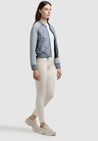 Kvinde iført lyseblå quiltet bomberjakke, hvid tætsiddende top, cremefarvede leggings og beige sneakers stående mod en ensfarvet baggrund.