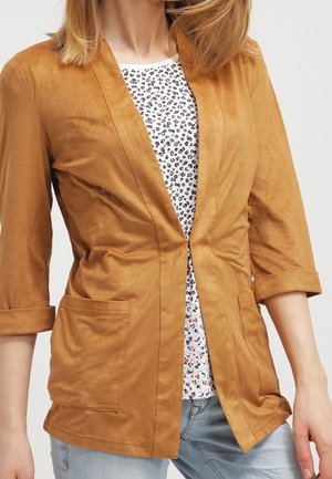 Frau trägt einen senfgelben, offen geschnittenen Blazer mit hochgekrempelten 3/4-Ärmeln über einem weißen Top mit Blumenmuster und hellblauen Jeans.