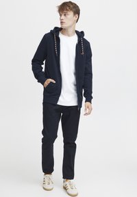 Marineblå zip-up hettegenser med hette og mønstrede snor, kombinert med en hvit t-skjorte, mørke bukser og hvite joggesko med beige striper.