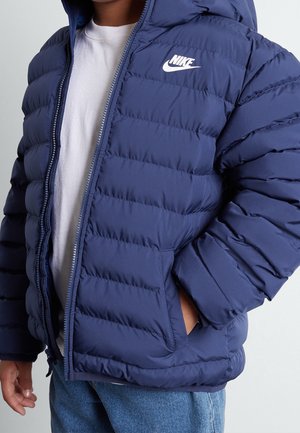 Dziecko w granatowej pikowanej kurtce Nike, na białej koszulce i niebieskich dżinsach, z rękami w kieszeniach kurtki.