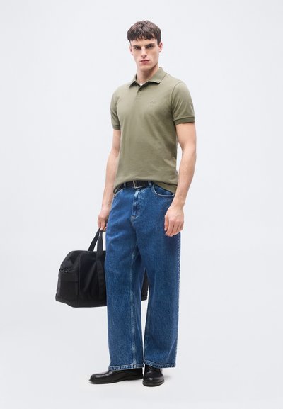 Jeune homme portant un polo vert olive, un jean bleu ample et des chaussures noires, tenant un sac de sport noir sur un fond uni.