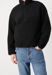 Maglione in pile nero con collo a mezza zip e superficie texturizzata, abbinato a jeans denim blu con vestibilità classica.