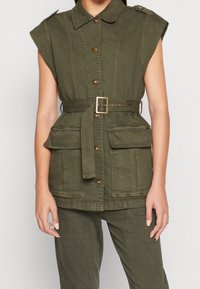 Gilet utility sans manches vert olive avec col, boutons dorés et taille ceinturée. Présente deux grandes poches avant et un design structuré.