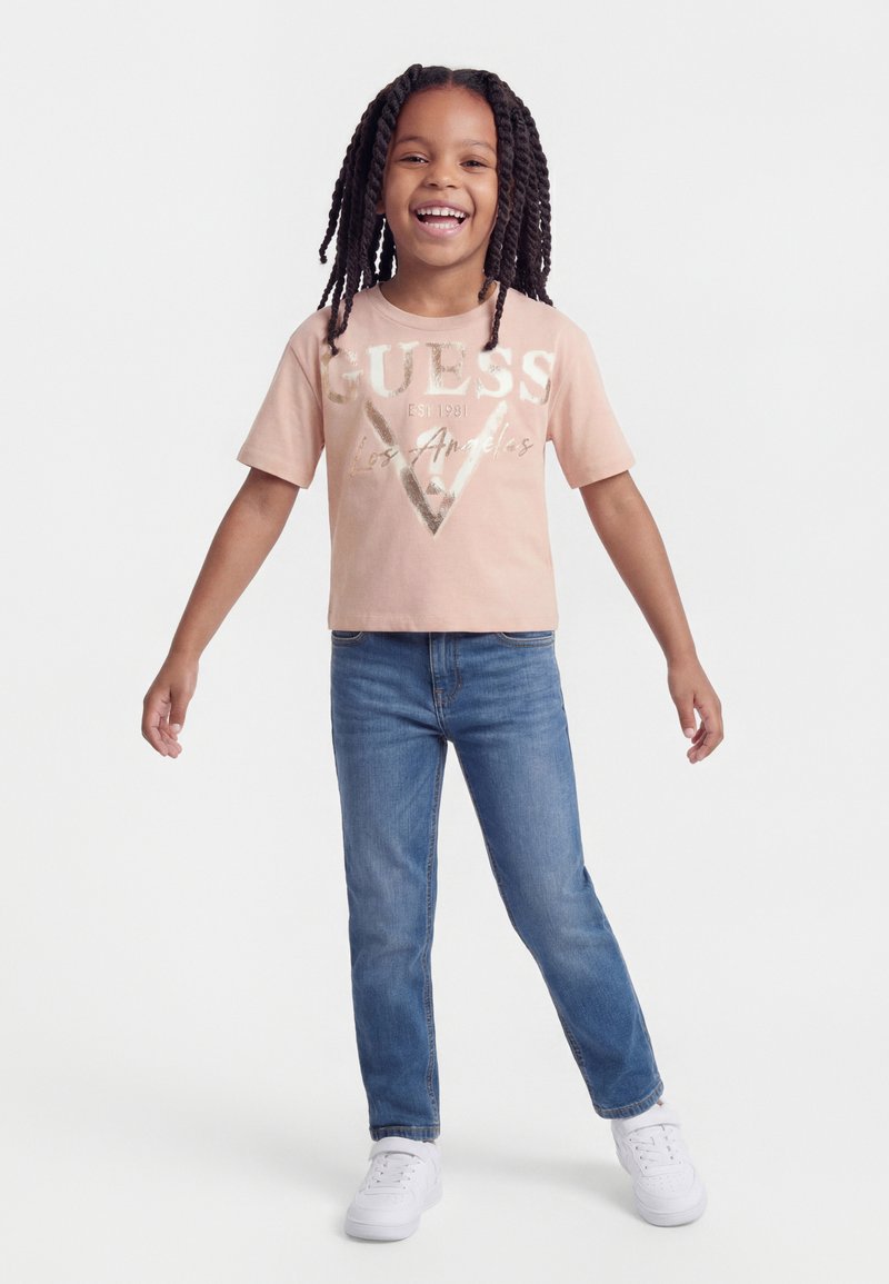 Enfant souriant avec de longs cheveux tressés, portant un T-shirt rose GUESS, un jean bleu et des baskets blanches, debout les bras légèrement écartés.