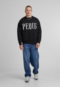 Svart genser med hvit "PEQUS" tekst, kombinert med løse blå jeans og sølvfargede joggesko. Stoffet ser mykt og strukturert ut.