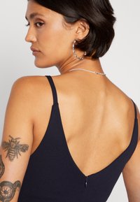 Navy plava haljina s tankim naramenicama, ima duboki V-izrez na leđima i mali detalj s patentnim zatvaračem. Glatka tkanina s mat teksturom.