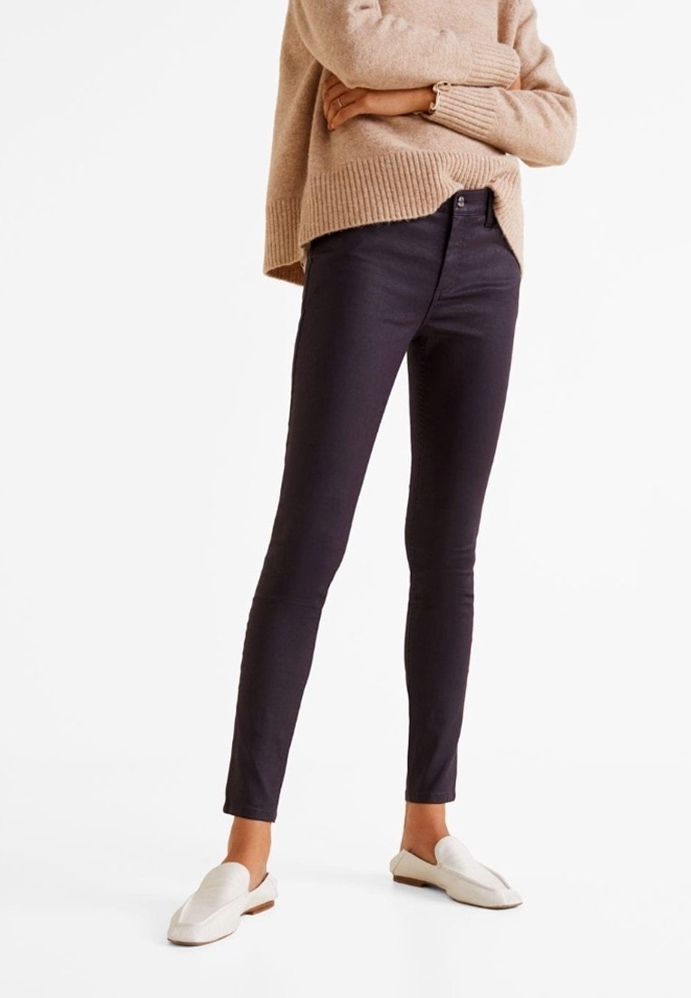 Personne portant un pantalon skinny noir, un pull en maille épaisse beige et des mocassins blancs sans lacets, debout les bras croisés sur fond blanc.