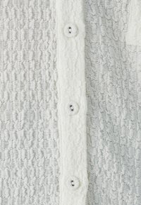 Texturovaná bílá tkanina s mačkaným vzorem; pět knoflíků vpředu; měkká a nadýchaná textura; knoflíkové dírky viditelné na knoflíkové liště.