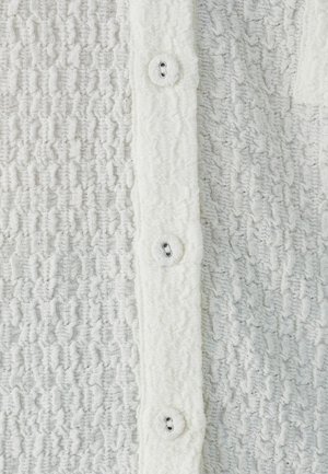 Texturovaná bílá tkanina s mačkaným vzorem; pět knoflíků vpředu; měkká a nadýchaná textura; knoflíkové dírky viditelné na knoflíkové liště.