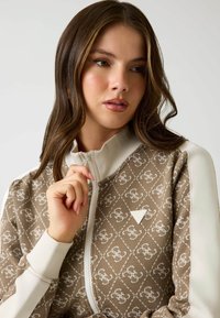 Braune Zip-Jacke mit einem gemusterten Design, weißen Akzenten und einem dreieckigen Logo. Verfügt über einen hohen Kragen und eine strukturierte Oberfläche.