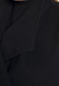 Manteau noir avec un col élégant, fabriqué en tissu texturé. Les détails en gros plan mettent en avant les coutures et les lignes épurées du design.
