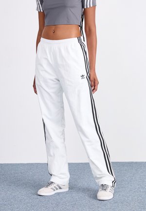Persona con pantalones de chándal blancos Adidas con rayas negras, top corto gris y zapatillas Adidas grises, de pie sobre una alfombra gris.