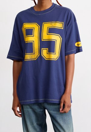 Camiseta oversize color azul marino hecha de algodón, con estampado desgastado en amarillo del número "95" en el frente y logo de Nike en la manga. Mangas cortas, ajuste casual.