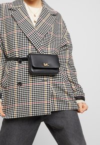 Czarny i biały płaszcz w kratę houndstooth z złotymi detalami. Dołączona czarna torba z paskiem z logo. Szare dżinsy i warstwowe naszyjniki dopełniają stylizację.