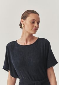 Abito blu navy in tessuto testurizzato con maniche corte, dotato di uno scollo rotondo e di una vita con nodo. Orecchini astratti dorati completano il look.