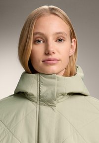 Mladá žena s blond vlasmi, oblečená v svetlozelenom prešívanom kabáte s vysokým golierom, na jednoduchom sivom pozadí.