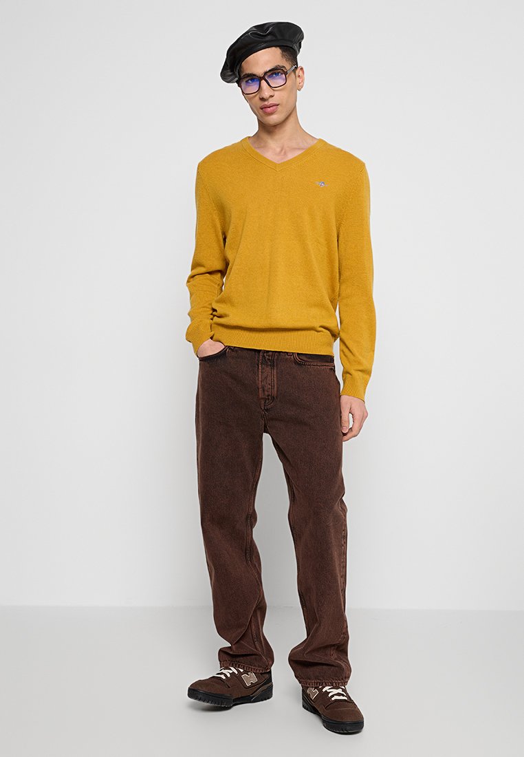 Gul v-ringad tröja, bruna kordfläskbyxor och bruna sneakers. Tillbehören inkluderar en svart basker och blå glasögon. Enkelt mönster, varma toner.