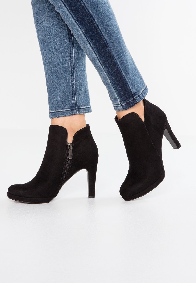 Paire de bottines en daim noir avec fermeture éclair latérale et talons hauts, portées avec un jean en denim bleu.