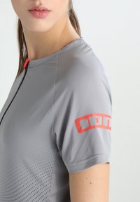 Camiseta atlética gris de manga corta, con cuello de media cremallera, tejido texturizado y logo en acento rojo en la manga. Presenta un diseño con patrón de puntos.