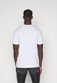 Fehér, rövid ujjú póló, pamutanyagból készült, kerek nyakkivágású, sima textúrájú, sötétszürke rövidnadrággal viselve, amelyen kisebb díszítések találhatók.