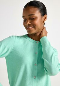 Ljusgrön kofta med rund halsringning, långa ärmar och fem silverknappar framtill. Mjuk textur, avslappnad design.
