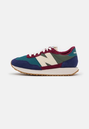 Mehrfarbiger New Balance Sneaker mit blauen, grünen, burgunderfarbenen und cremefarbenen Paneelen, weißen Schnürsenkeln und einer Gummilaufsohle, von der Seite gezeigt.