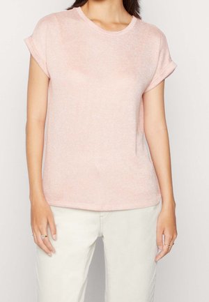 Vrouw draagt een lichtroze, gebreide top met korte mouwen en opgerolde manchetten, gecombineerd met roomwitte broek, staand tegen een neutrale achtergrond.