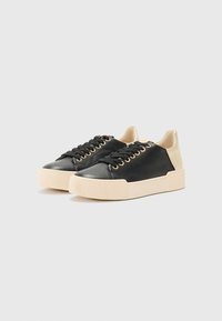 Black leather sneakers with gold eyelets, lace-up design, contrasting beige heel, and textured beige rubber sole. Flat, casual style.