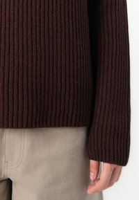 Nahaufnahme einer Hand neben beigen Hosen, die einen dunkelbraunen, grob gestrickten Pullover mit langen Ärmeln trägt.