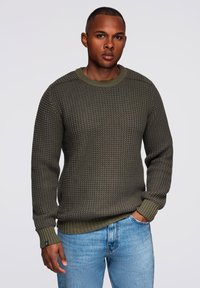 Suéter de punto verde oliva con un patrón texturizado, cuello redondo y puños acanalados, combinado con jeans azul claro. Diseño simple y casual.