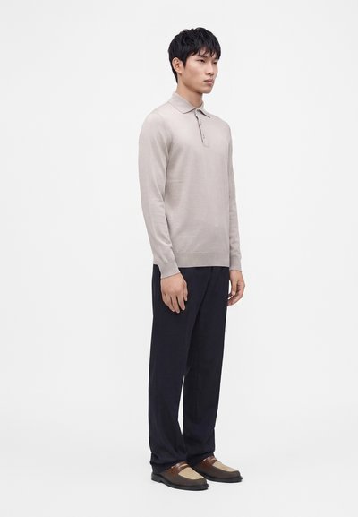 Homme debout portant un polo beige clair à manches longues, un pantalon ample foncé et des mocassins marron bicolores sur fond blanc.