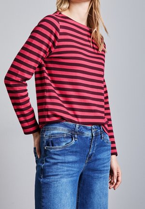 Vrouw die een rood-zwart horizontaal gestreept shirt met lange mouwen draagt, ingestopt in blauwe spijkerbroek, staand tegen een effen achtergrond.