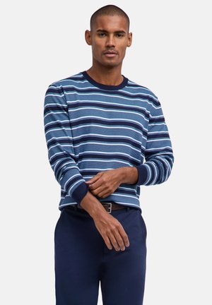 Uomo che sistema il polsino di un maglione a righe blu navy e azzurro, indossando pantaloni blu scuro e una cintura marrone, contro uno sfondo chiaro e uniforme.