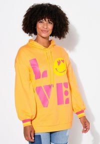 Felpa oversize gialla brillante con testo "LOVE" rosa e emoji sorridente, dotata di cordoncini, polsini a costine e una texture accogliente.