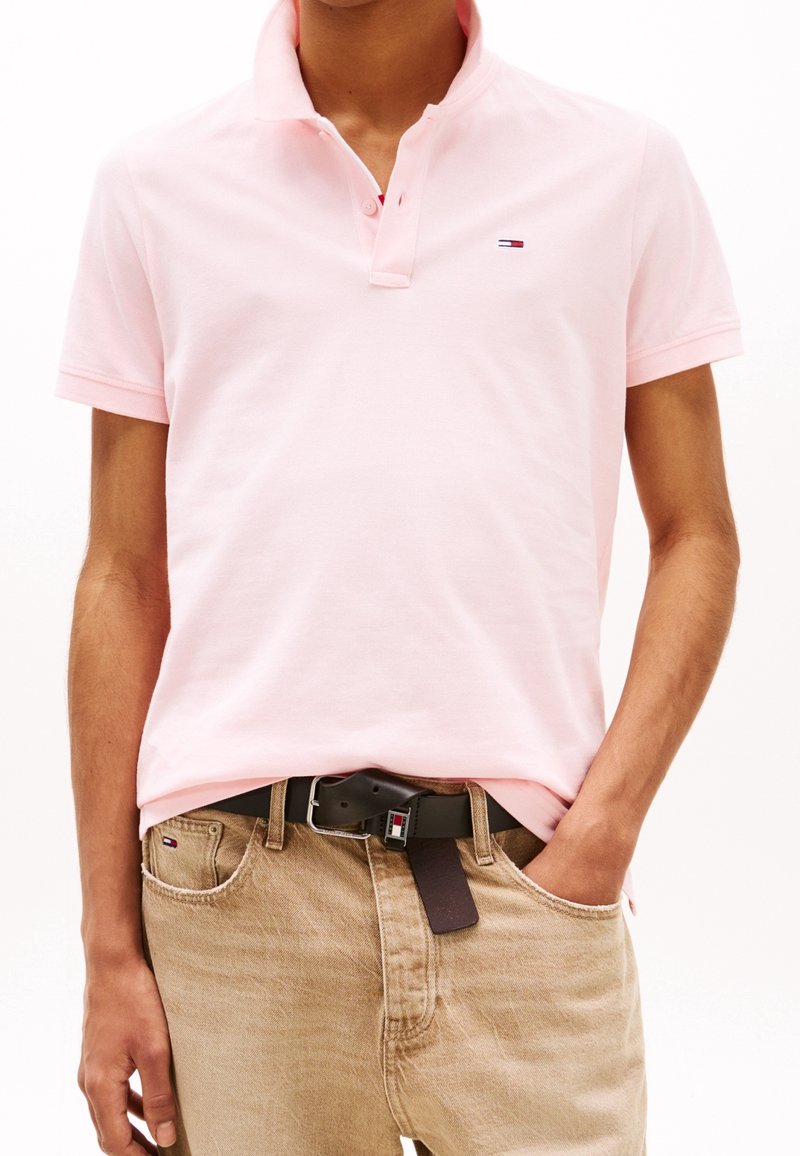 Polo rose avec un col classique et deux boutons, associé à un jean beige et une ceinture noire avec un petit logo multicolore.