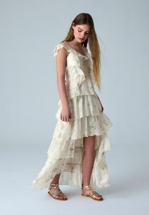 Junge Frau steht und trägt ein mehrlagiges, cremefarbenes Blumenkleid mit Rüschen und metallische Gladiator-Sandalen, den Kopf geneigt.