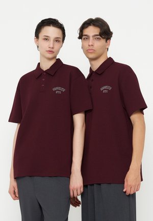 Deux personnes portant des polos rouge foncé assortis avec le texte "Brooklyn NYC", debout côte à côte sur un fond uni.