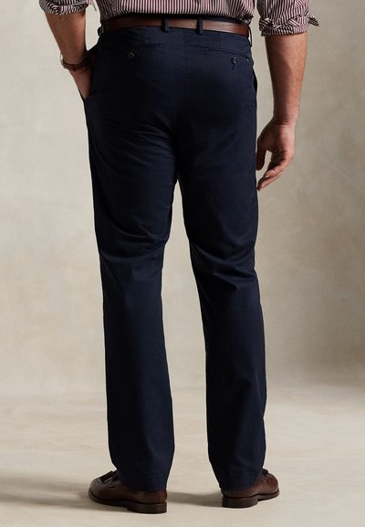 Egy férfi tengerészkék nadrágot, barna övet, barna alkalmi cipőt és csíkos inget visel, amelynek fel van hajtva az ujja, és az egyik kezét a zsebében tartja.