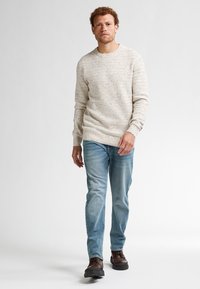 Crème-kleurige, getextureerde gebreide trui met een ronde hals, gecombineerd met lichtblauwe denimjeans en donkerbruine schoenen. Model aan het lopen.