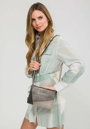 Donna con lunghi capelli biondi che indossa un abito pastello chiaro con cintura, che tiene una piccola borsa a tracolla grigia con dettagli metallici.