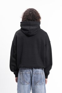 Personne avec des cheveux bouclés portant un sweat à capuche noir et un jean léger bleu clair, se tenant de dos à la caméra sur un fond blanc.