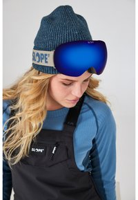 SLOPE-märket skidglasögon med blå spegelglas, som bärs över en stickad blå mössa. Modellen har också på sig en svart hängseloverall med logodetalj.