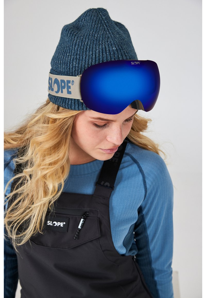 SLOPE-märket skidglasögon med blå spegelglas, som bärs över en stickad blå mössa. Modellen har också på sig en svart hängseloverall med logodetalj.