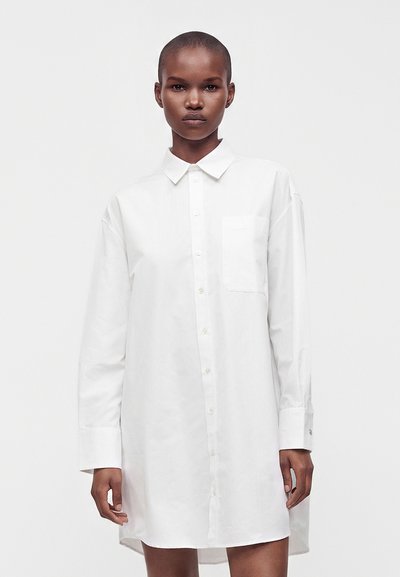 Femme aux cheveux courts portant une longue robe chemise blanche à boutons avec un col et une poche poitrine sur un fond clair uni.