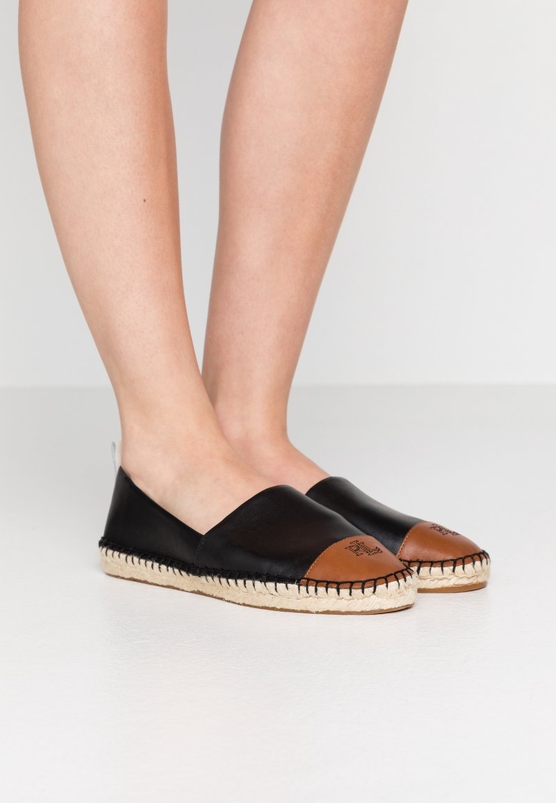 Pieds portant des espadrilles en cuir noir et marron avec des semelles en jute cousues sur un sol blanc et un fond uni.